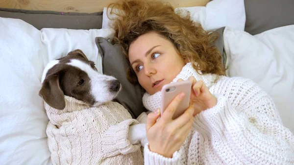 Kadın köpeğine yatakta uzanmış akıllı telefonu nasıl kullanacağını öğretiyor.. — Stok fotoğraf