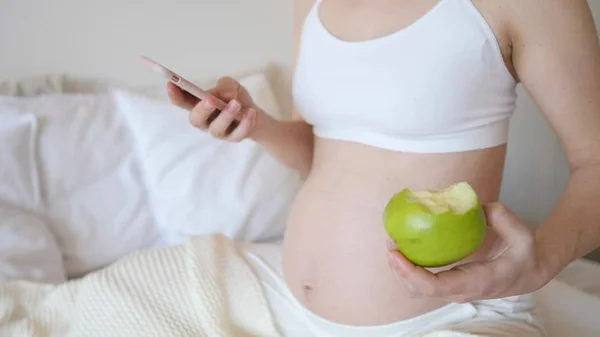 Fechar-se de mulher grávida usando Smartphone e comer maçã verde . — Fotografia de Stock