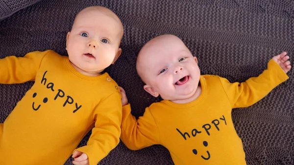 Zwillingsbabys tragen fröhliche ockerfarbene T-Shirts und liegen auf einer Strickdecke. Ansicht von oben. — Stockfoto