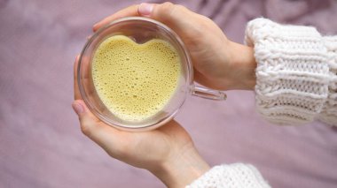 Altın sarısı badem latte kalp şeklinde cam bardakta tutulan kadın elleri.