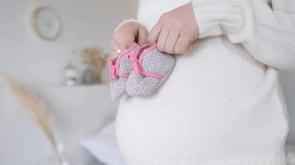 Gravid kvinna som håller små skor för det ofödda barnet på magen. Närbild. — Stockfoto