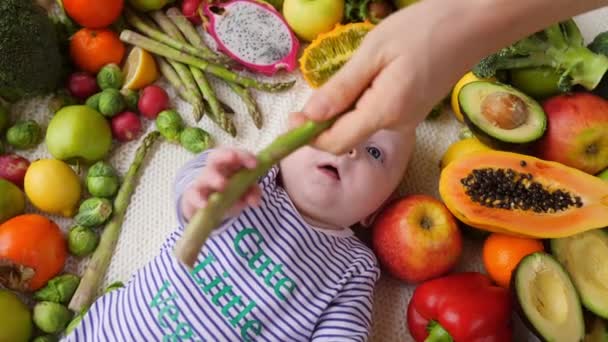 Vegano bebé sosteniendo espárragos acostado en verduras y frutas . — Vídeo de stock