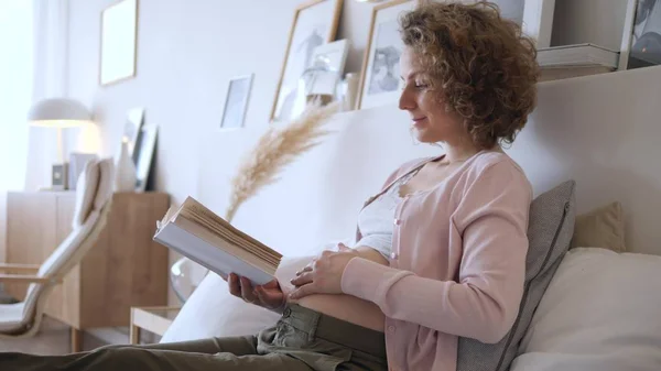 Беременность, отдых, люди и ожидания Концепция - счастливая беременная женщина лежит в постели с книгой дома . — стоковое фото