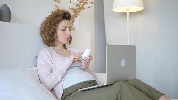 Беременная женщина с ноутбуком и чтение этикетки на бутылке лекарств дома — стоковое фото