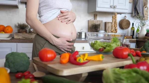Nahaufnahme einer schwangeren Frau, die ihren Bauch beim Kochen einer gesunden Mahlzeit hält. — Stockfoto