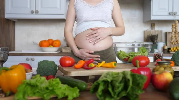 Gravid kvinna matlagning färsk sallad och hålla sin mage på kök. — Stockfoto