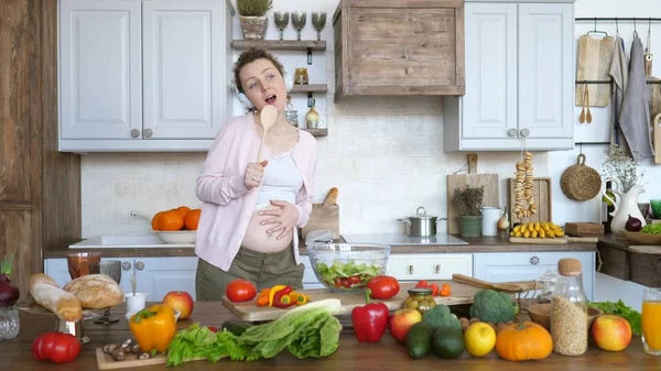 Счастливая концепция беременности. Веселая беременная девушка танцует и поет на кухне . — стоковое фото