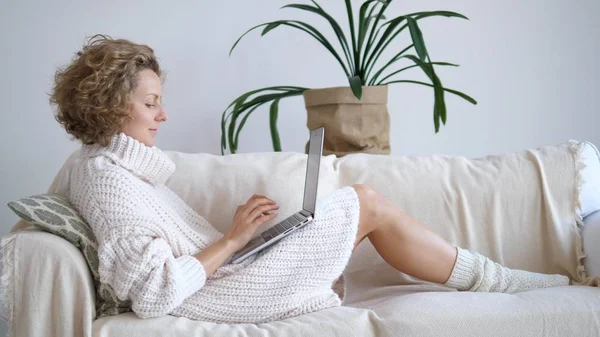Молодая женщина в трикотажном свитере с помощью ноутбука дома отдыхает на диване — стоковое фото