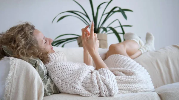 Ung kvinna i stickad tröja med hjälp av smartphone liggande på soffan — Stockfoto