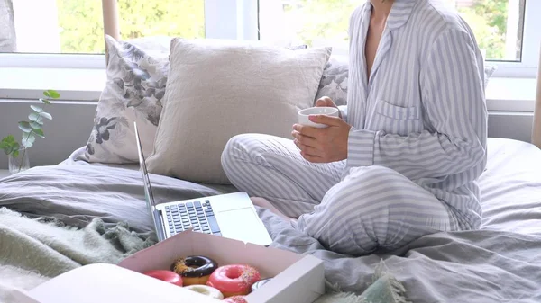 Женщина в пижаме с ноутбуком на удобной кровати с чашкой кофе — стоковое фото