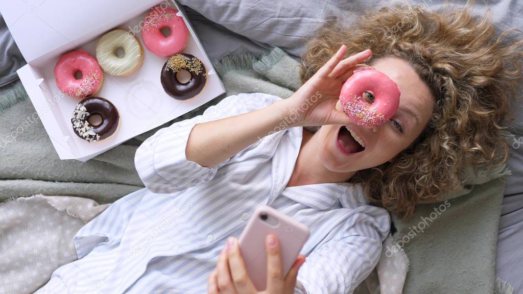 Young Woman Taking Selfie In Bed With Doughnuts