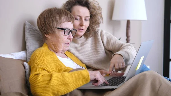 Barnebarn som hjelper bestemor med laptop liggende i sengen. Teknologi, mennesker og generasjonskonsept . – stockfoto