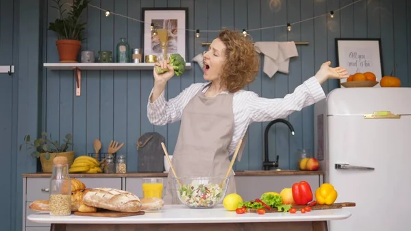 Ung glad kvinna sjunger och dansar på köket medan matlagning hälsosam mat. — Stockfoto