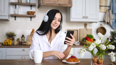 Evdeki mutfakta cep telefonu kullanan esmer bir kadın. Kulaklık takıyor.