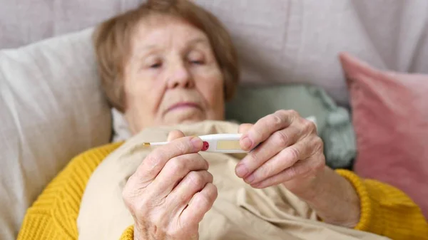 Mujer mayor enferma revisando termómetro en la cama en casa —  Fotos de Stock