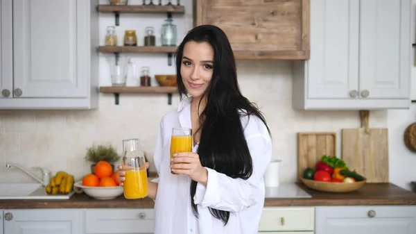 Mulher morena bonita derramando suco de laranja em vidro na cozinha pela manhã — Fotografia de Stock