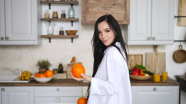 Jovem morena mulher segurando laranjas na cozinha e sorrindo . — Fotografia de Stock