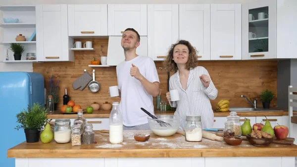 Casal jovem com copos de café dançando juntos na cozinha enquanto cozinha café da manhã . — Fotografia de Stock