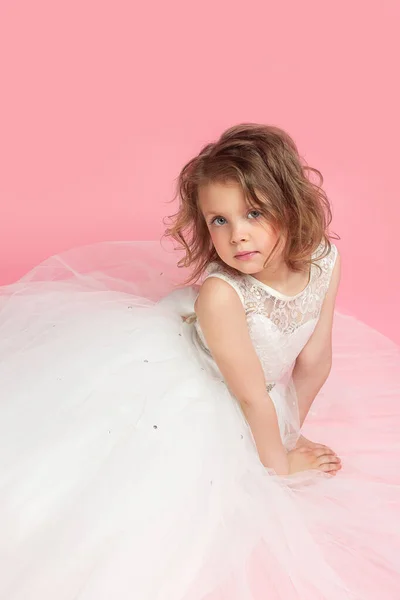 Portrait of a cute little blonde girl in a beautiful delicate white dress isolated on a pink background. The girl looks into the frame with a smile. Space for text. On pink background with copy space