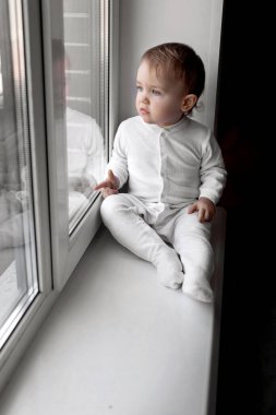 Beyaz tulumlu küçük bir çocuk evin pencere eşiğinde oturur ve pencereden dışarı bakar. Karantina ve kendini izole etme