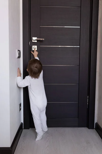 Küçük bir çocuk odadan çıkmaya çalışıyor. Beyaz tulumlu bir bebek kapıda dikiliyor. Ev güvenliği. Küçük bebek odadan kaçmak için kapı koluna ulaşmaya çalışıyor.