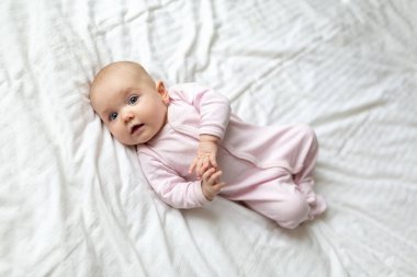 Küçük şirin bir bebek, ebeveyn yatak odasındaki beyaz bir çarşafın üzerinde yatarken gülümsüyor. Yeni doğan bebeğin yakın plan portresi kameraya bakıyor.