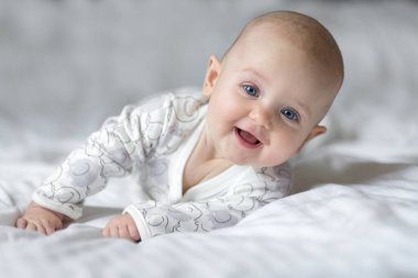 Küçük şirin bir bebek, ebeveyn yatak odasındaki beyaz bir çarşafın üzerinde yatarken gülümsüyor. Yeni doğan bebeğin yakın plan portresi kameraya bakıyor.