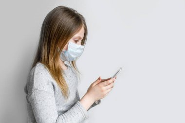 Hijyenik koruyucu maskeli küçük bir kız enfeksiyonu önlemek için elinde telefon tutuyor. Bulaşıcı hastalıklara karşı koruma, koronavirüs. Sağlık ve Tıp Konsepti.