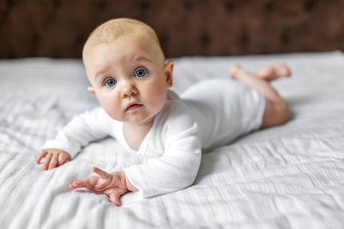 6 aylık beyaz bir tulumla evde kameraya bakan tatlı bir bebek. Bir erkek bebeğin battaniyeye uzanmış portresi. Mutlu annelik çocukluğu. Avrupa görünümlü küçük bir çocuk