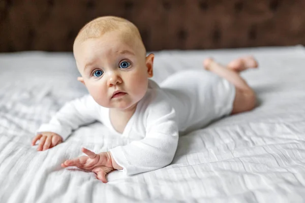 6 aylık beyaz bir tulumla evde kameraya bakan tatlı bir bebek. Bir erkek bebeğin battaniyeye uzanmış portresi. Mutlu annelik çocukluğu. Avrupa görünümlü küçük bir çocuk