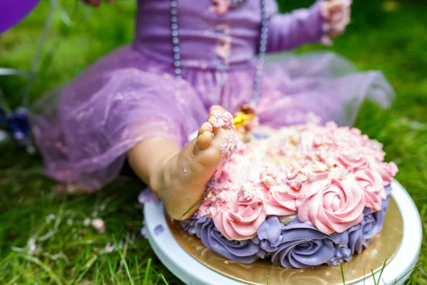 Close-up child breaks the cake on the green grass in the Park. A sweet pie on her feet. Crash pink cake at the celebration of the first birthday of the girl. One year old baby. Smash cake.
