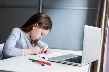 Okul çağındaki kız çalışmak için aletler kullanıyor. Küçük kız evde dizüstü bilgisayarıyla ödevini yapıyor. Çocuklar için eğitim ve uzak mesafe eğitimi. Karantina sırasında evde eğitim. Evde kalıp eğlenin.