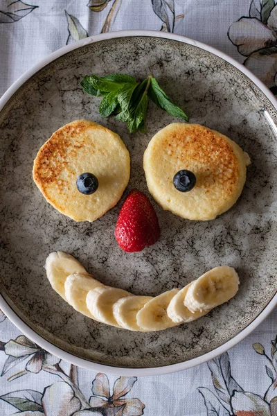 Healthy breakfast. Funny kid breakfast in the shape of face. Cooking and serving food at home Pancakes with blueberries, strawberries and banana. Vegitarian dish with berries and fruits. Creative food