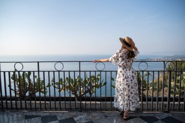 Hasır şapkalı, beyaz elbiseli şık bir kız balkonda duruyor. Tatil için uygun kıyafet. Güzel siyah saçlı bir kız denize hayrandır. Modaya uygun yaz tarzı..