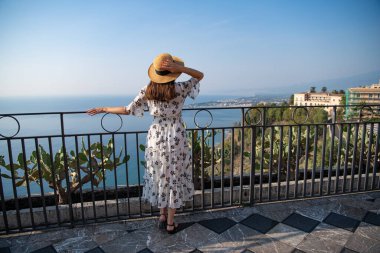 Hasır şapkalı, beyaz elbiseli şık bir kız balkonda duruyor. Tatil için uygun kıyafet. Güzel siyah saçlı bir kız denize hayrandır. Modaya uygun yaz tarzı..