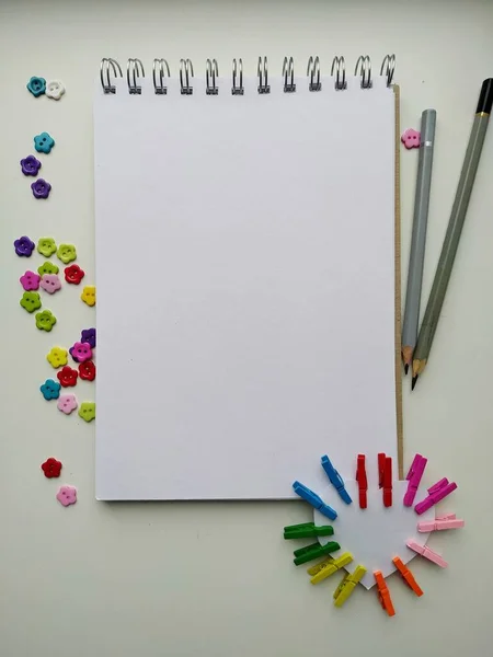Cuaderno para dibujar sobre un fondo blanco con botones multicolores en forma de flores y pinzas de ropa de colores —  Fotos de Stock