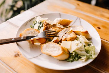 Güzel bir tabakta Sezar salatası güneşli bir günde bir kafede ahşap bir masanın üzerinde duruyor. Sezar, tavuk, beyaz ekmek, parmesan ve ançüez soslu. Tabakta çatal ve bıçak var, arkasında da saksılarda yeşil bitkiler..