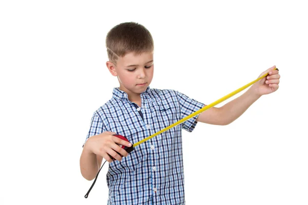 Een kleine knappe bouwer jongen in blauwe geruite shirt kijkt naar een meetlint, geïsoleerd op witte achtergrond. — Stockfoto