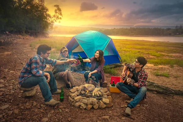Happy friends camping party playing music and enjoying bonfire in nature and lake, Traveler camping on the river/mountain. With a relaxing drink in the sunset. The concept of hiking/camping and relax.