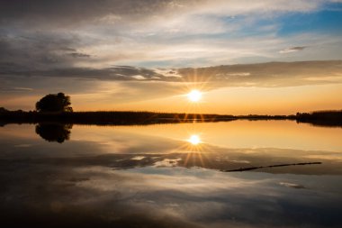 Temiz ve güzel bir gölün huzurlu yüzeyinde ılık bir bahar akşamında güzel bir günbatımı. Kristal suda, parlak güneşi sıcak ışınlarla yansıtır. Gri ve beyaz bulutlar gökyüzünde süzülür
