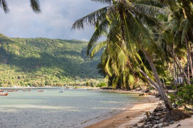 Tropikal adadaki güzel deniz lagünü ve kumlu plaj manzarası. Dalgalarda uzun balıkçı tekneleri sallanıyor, denize giden gece çıkışını bekliyorlar. Güneş fırtına bulutlarının arasından geçiyor..