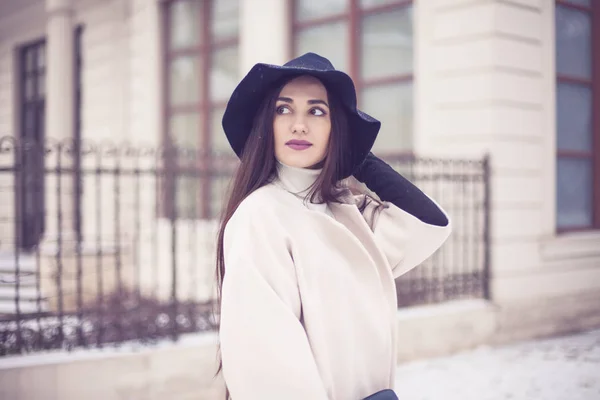 Charming young woman in coat with long brunette hair wearing a hat enjoying snowfall in big city. Cheerful emotions, smiling, christmas mood, positive face emotions, winter weather. Leather gloves and stylish purse. — Stock Photo, Image