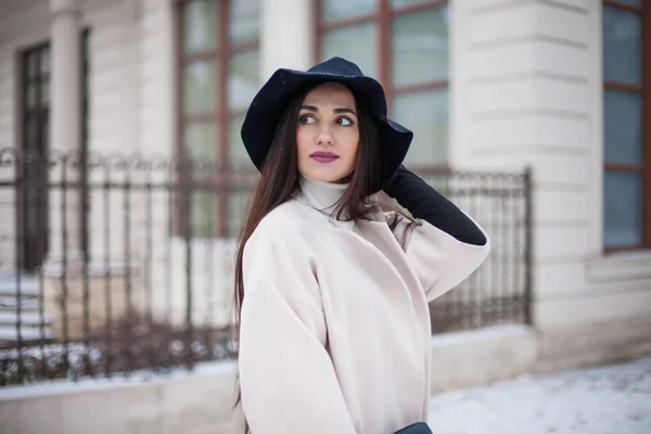 Очаровательная молодая женщина в пальто с длинными брюнетками в шляпе, наслаждающаяся снегопадом в большом городе. Веселые эмоции, улыбка, рождественское настроение, позитивные эмоции лица, зимняя погода. Кожаные перчатки и стильная сумочка . — стоковое фото