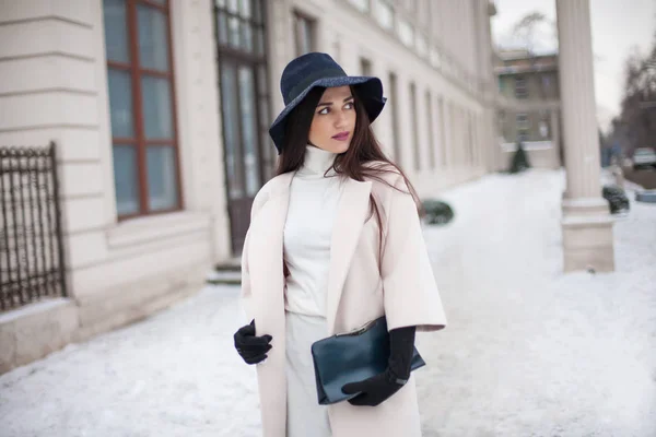 Очаровательная молодая женщина в пальто с длинными брюнетками в шляпе, наслаждающаяся снегопадом в большом городе. Веселые эмоции, улыбка, рождественское настроение, позитивные эмоции лица, зимняя погода. Кожаные перчатки и стильная сумочка . — стоковое фото