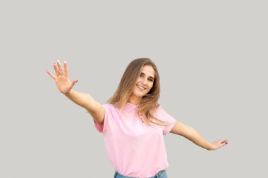 Gülümseyen ve beyaz dişli genç bir kız. Ellerini yana doğru sallıyor. Kameraya bak. Pembe tişört ve kot pantolon giymişti. Gri arkaplanda izole.