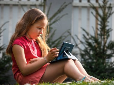 Covid-19 virüsünün karantina döneminde uzaktan öğrenme. Uzun saçlı sevimli küçük kız evden ders çalışıyor, bahçede çimlerin üzerinde oturuyor. İnternet üzerinden bir tablet ve uzaktan çalışma kullanır.