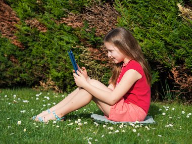 Covid-19 virüsünün karantina döneminde uzaktan öğrenme. Uzun saçlı sevimli küçük kız evden ders çalışıyor, bahçede çimlerin üzerinde oturuyor. İnternet üzerinden bir tablet ve uzaktan çalışma kullanır.