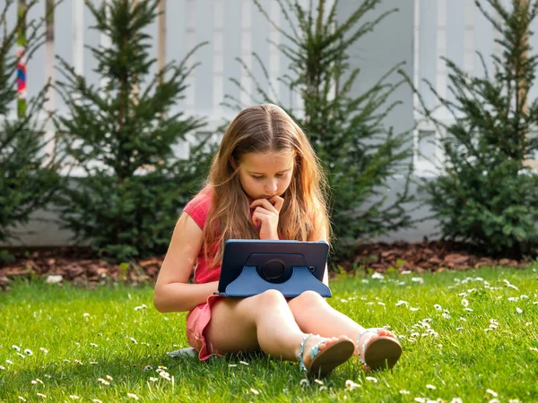 Distance learning during the quarantine period for the virus Covid-19. Cute little schoolgirl with long hair is studying from home, sitting in the garden on the grass. Uses a tablet and remote work via the Internet.