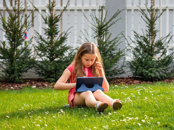 Distance learning during the quarantine period for the virus Covid-19. Cute little schoolgirl with long hair is studying from home, sitting in the garden on the grass. Uses a tablet and remote work via the Internet.