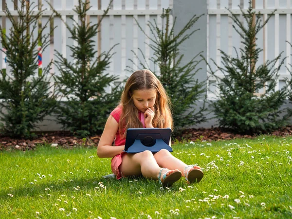 ウイルスCovid 19の隔離期間中の距離学習 長い髪をしたかわいい女の子は家から勉強していて 芝生の上の庭に座っています インターネットを介してタブレットやリモートワークを使用しています — ストック写真